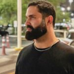 Plano fechado do rosto de perfil do paciente da Dra. Vanessa Melo Bucomaxilofacial - Homem branco de barba feita e cabelo baixo, de preto. sorridente.