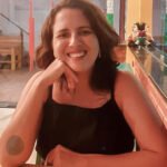 Plano médio da paciente da Dra. Vanessa Melo Bucomaxilofacial - Mulher branca sorridente sentada e apoiada com os braços na mesa com uma mao no queixo, vestida de preto e com uma tatuagem circular no cotovelo.