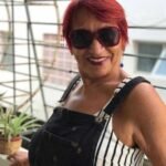 Plano médio da paciente da Dra. Vanessa Melo Bucomaxilofacial - Idosa, jovial, Mulher preta de cabelos curtos, lisos e vermelhos, de óculos escuro grande e sorridente.