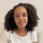 Plano médio da paciente da Dra. Vanessa Melo Bucomaxilofacial - Jovem, Mulher preta de cabelos grandes, volumosos, escuros e crespos, de óculos de grau grande e fino. camisa branca e funco branco.