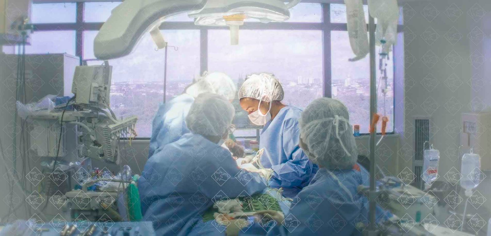 Plano aberto em destaque Dra. Vanessa Melo Bucomaxilofacial em ciruirgia no bloco de touca e máscara branca e Scrubs azul claro.