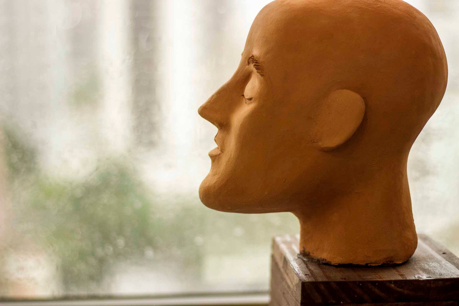 plano fechado proximo a uma janela uma escultura de barro de uma cabeça careca com o queixo destacado, perfil classe 3